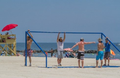 Open beach near blue water with five people standing near a soccer goal net