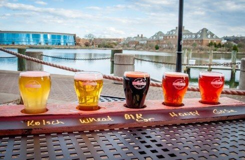 Outside patio table near water holding a flight tray of beers of various colors each with a name