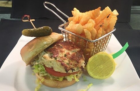 Grilled crabcake on a bun with lettuce and tomato next to a basket of fries on a white square plate