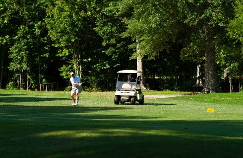 Golfer in shorts mid-swing out on a wooded golf course with a cart nearby