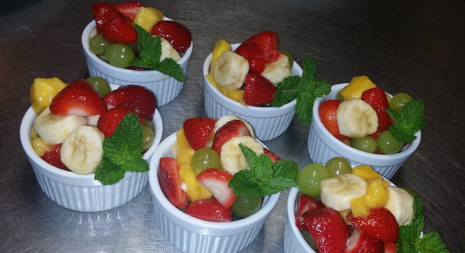 Six white ramekins filled with mixed fruit and min leaves on a silver countertop