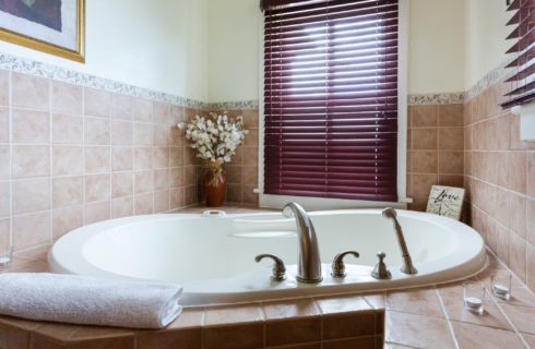 Large soaker tub in the corner of a bathroom with brown tile and a two windows with burgundy shades