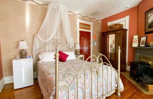 Four-poster brass-style bed with net canopy in room with fireplace, armoire and small fridge