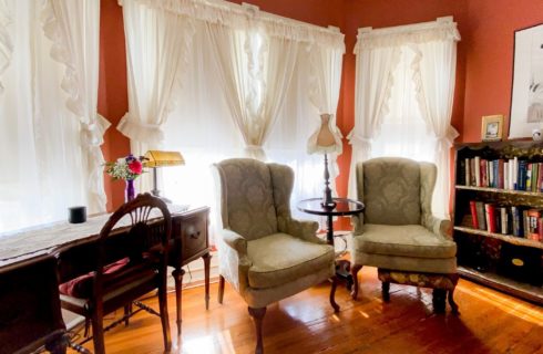 Sitting area with two wing-back chairs, writing desk and bookshelf in front of bright windows with white curtains