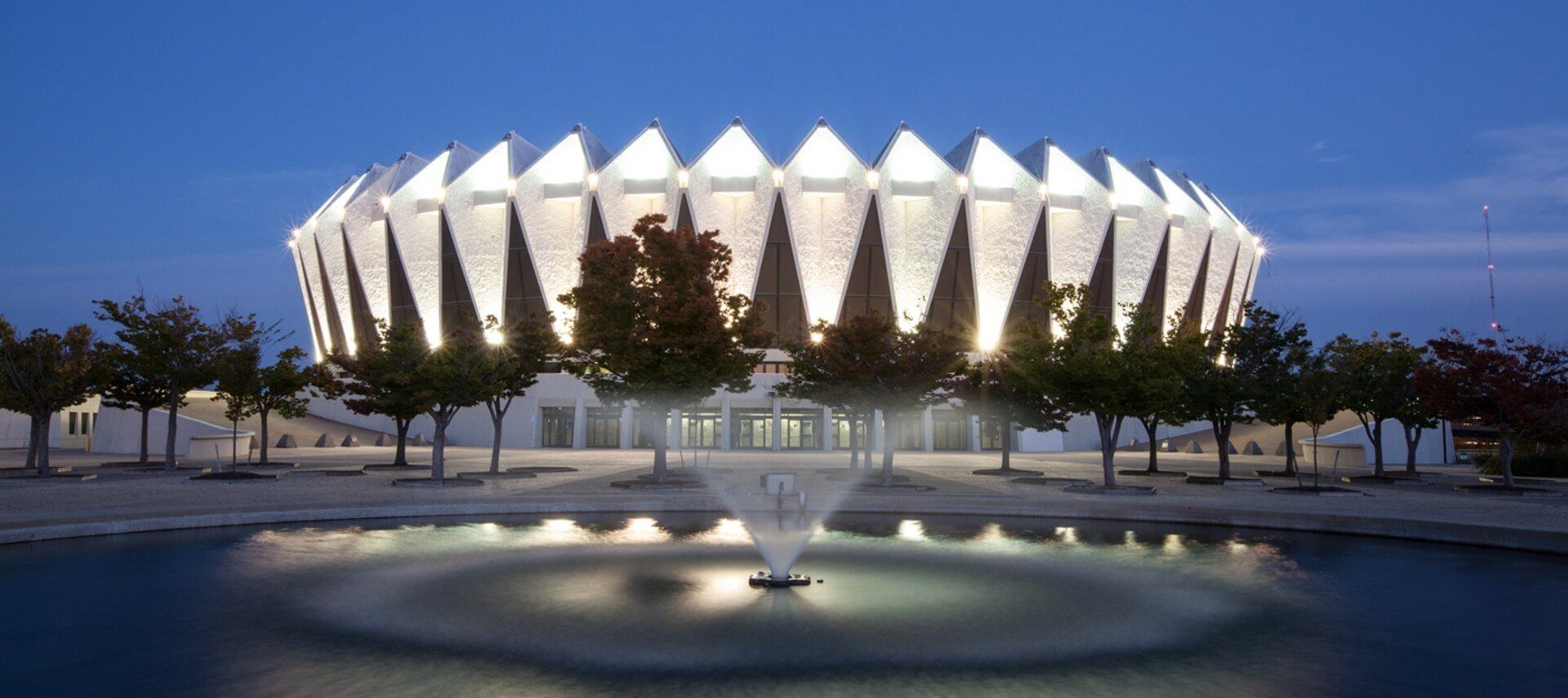 Coliseum style building lit up at night with rows of trees and a fountain in front