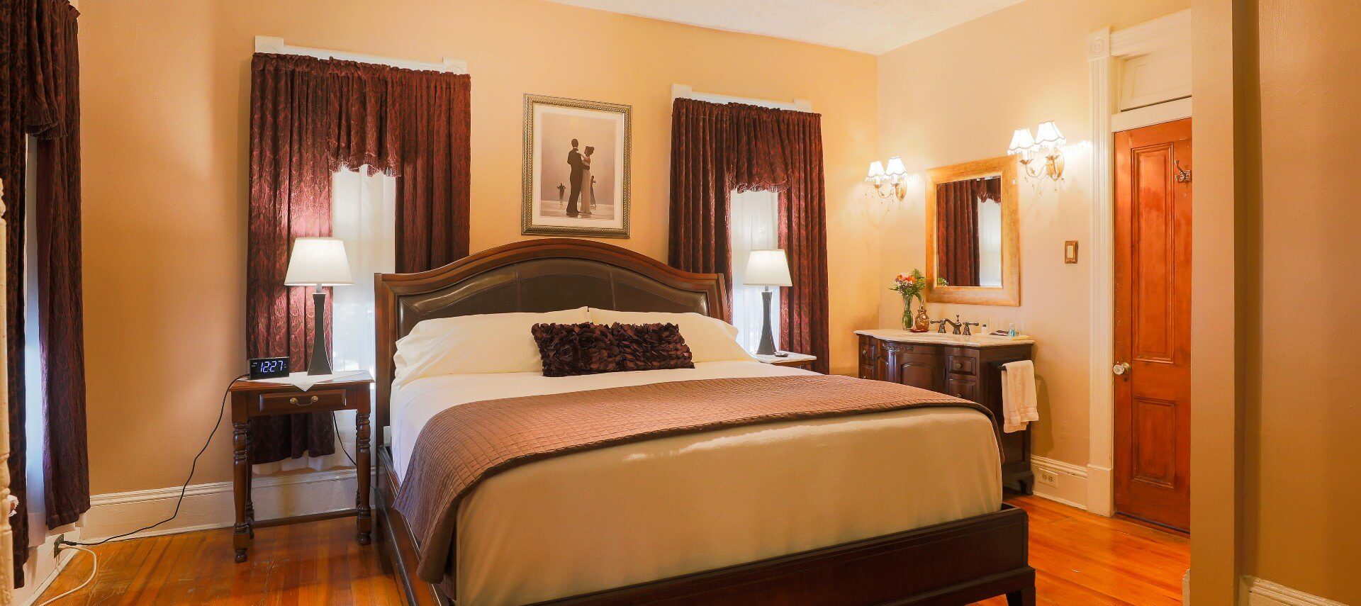 King bed in luxurious linens in front of two large windows and vanity with sink under a gold framed mirror