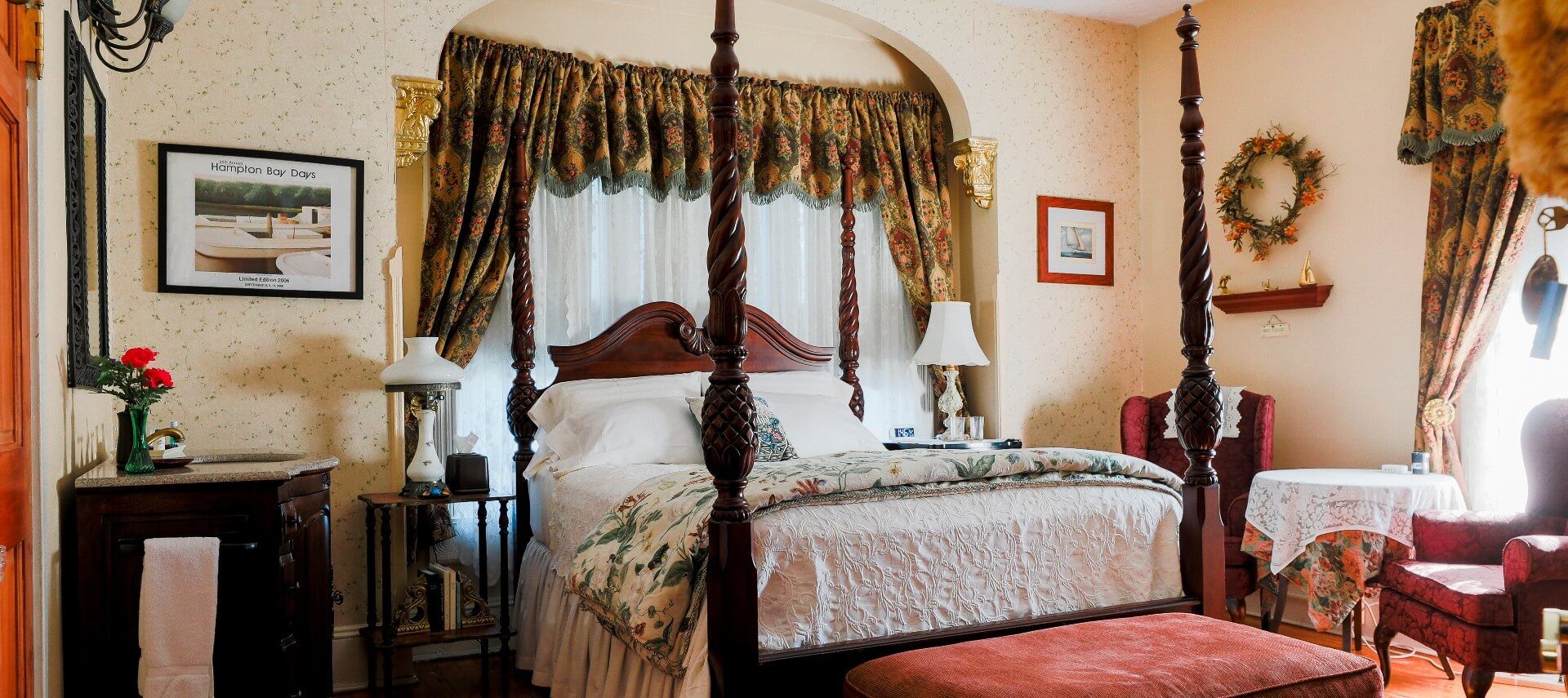 A four-poster cherry wood bed in front of large window in guest room with two sitting chairs and vanity with sink