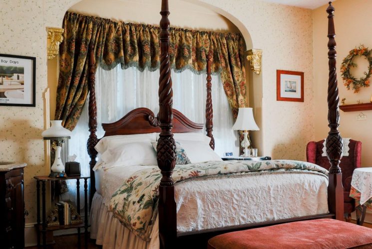 A four-poster cherry wood bed in front of large window in guest room with two sitting chairs and vanity with sink