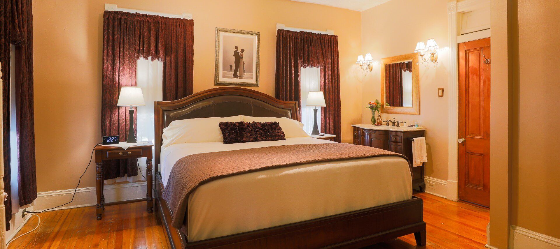 Elegant guest room with king bed, bedside tables, three windows and vanity with sink below gold framed mirror