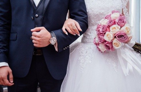 Bride in white with bouquet of white and pink flowers arm in arm next to a groom in a black suit