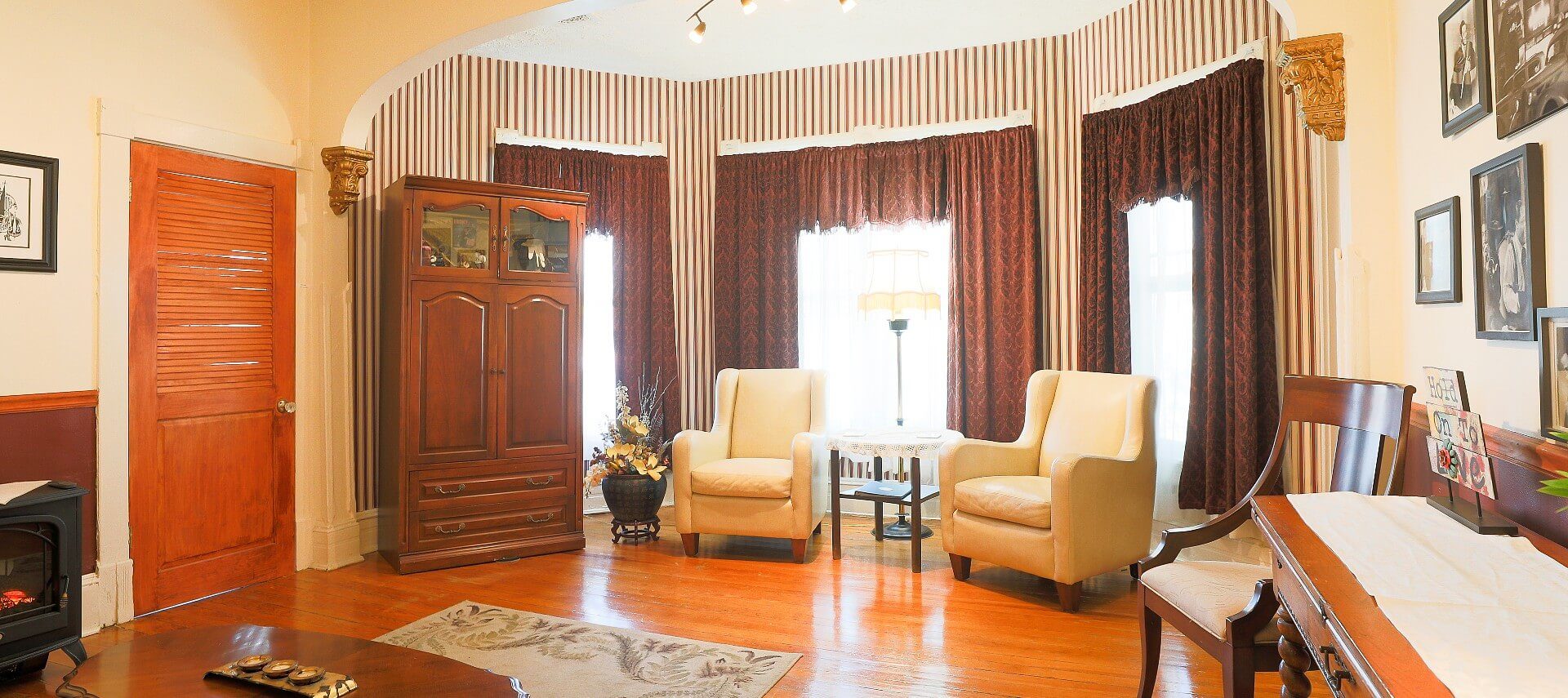 Large bay window with brown curtains and striped wallpaper behind two sitting chairs, side table, and tall armoire
