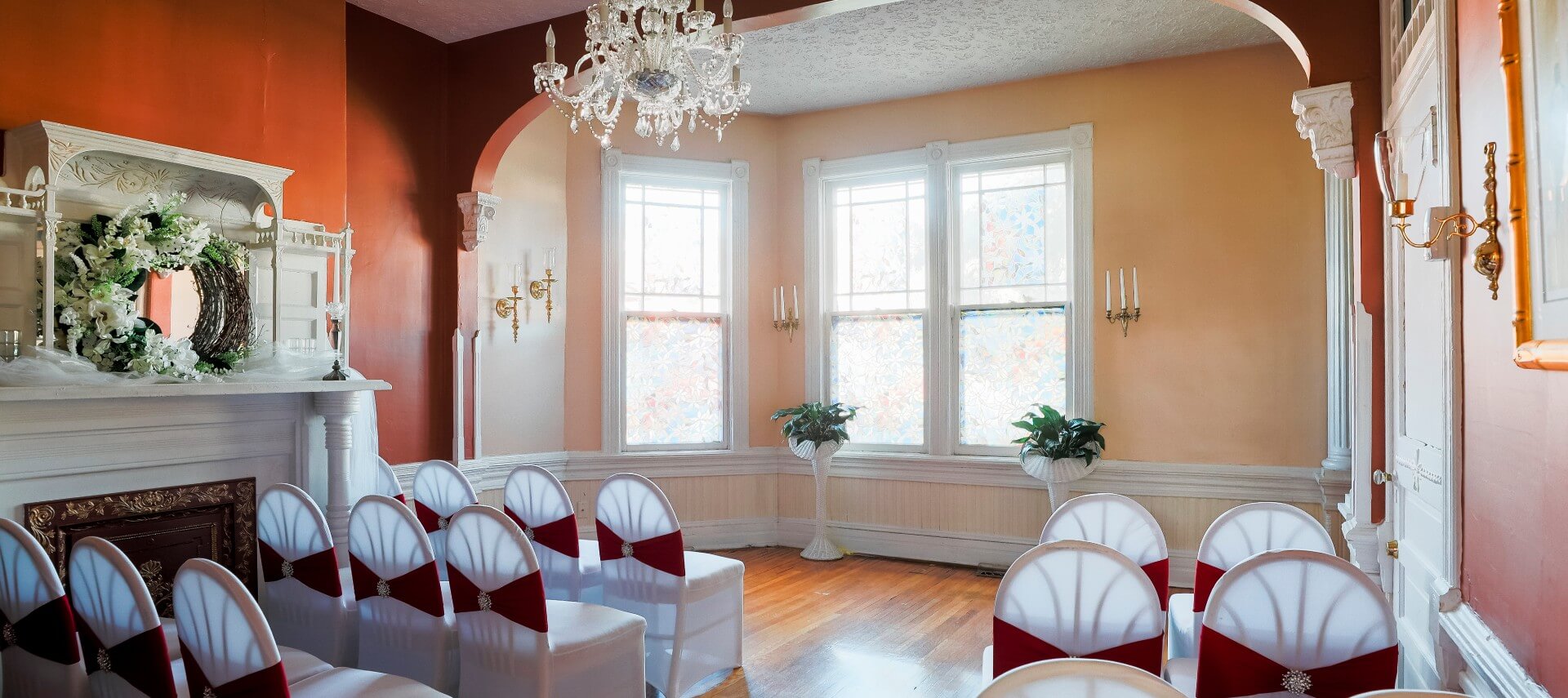 Large room in a home set up for a wedding with white chairs with red ribbons, white fireplace and large open windows