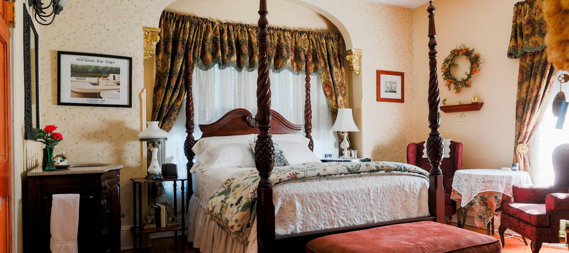 Four poster bed in guest room with wallpapered walls, dark brown vanity with sink and two upholstered wingback chairs